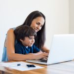 Young mother and son sitting at table and using laptop at home.