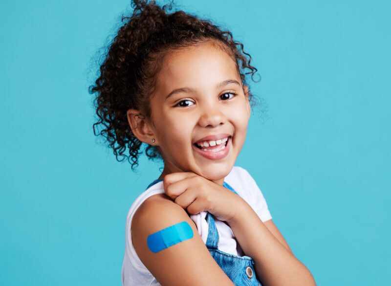  An elementary-aged girl smiling and showing off her bright blue bandaid on her arm after receiving a shot. 