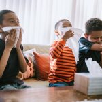 Three boys are sick at home, coughing and sneezing into tissues. They rest on the couch while they recover from their illness.