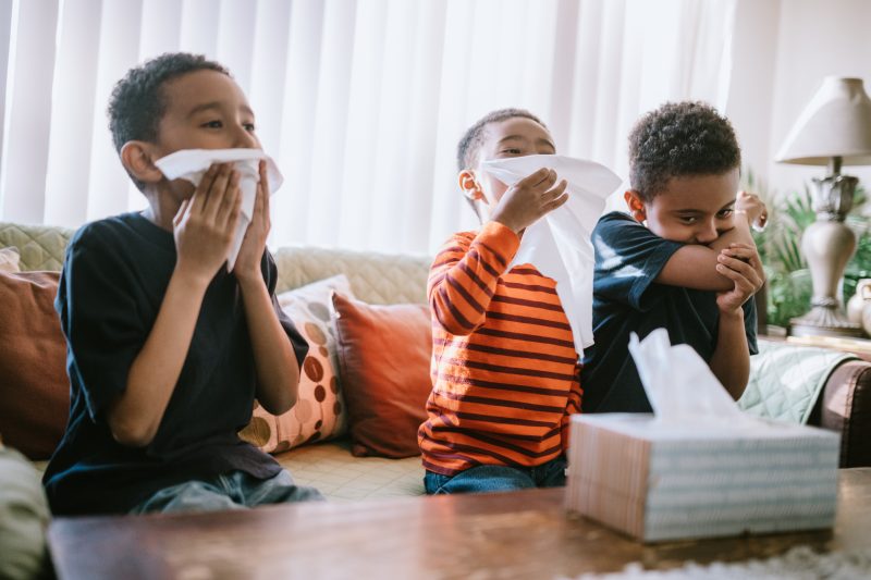 Three boys are sick at home, coughing and sneezing into tissues. They rest on the couch while they recover from their illness.