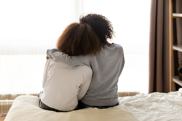 Rear back view black mother and daughter embrace sitting on bed at home.