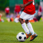 How to choose extracurricular activities for your kids African American Boy in Junior Football Team Leading Ball on Grass Training Field. Youth Soccer Player Kicking Ball