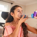 Asthma 101: Make a plan A female child sitting on a chair uses an asthma inhaler with a spacer. The room is well-lit, featuring large windows with sheer curtains and some indoor plants. The child wears a pink top.