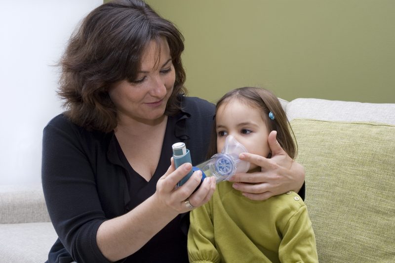 A mother helps a female child use an inhaler while sitting on a couch.
