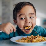 A front view of a hungry mixed-race boy eating fried rice for lunch with a spoon. The background is blurred.