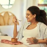 Pregnant, eating and woman with a smile, wellness and cheerful for nutrition, health and happiness at home. Pregnancy, female person and lady with joy, enjoying a snack and body care in living room