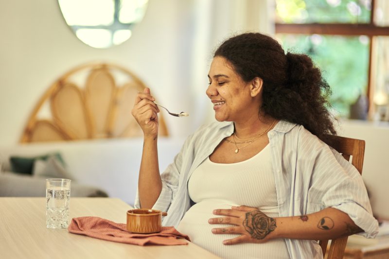 Pregnant, eating and woman with a smile, wellness and cheerful for nutrition, health and happiness at home. Pregnancy, female person and lady with joy, enjoying a snack and body care in living room