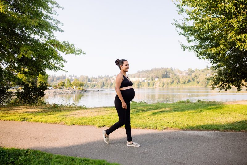 Pregnant woman of color taking a walk in athletic wear during summer next to a lake on a paved walking path with headphones in. 