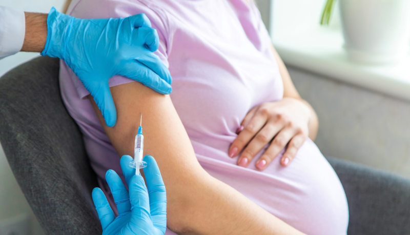 A pregnant woman is getting a vaccination. She lays her left hand gently on top of her stomach. A doctor with gloves on is holding her arm with one hand and holding a shot to her arm with the other. The image is cropped, so you only see the woman's body from neck to waist.