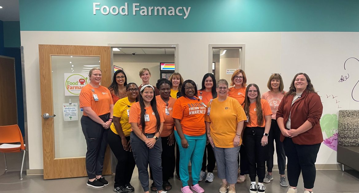 The Food Farmacy team standing together and smiling in front of a door labeled "Food Farmacy" at Akron Children's. They are wearing orange shirts and name badges.