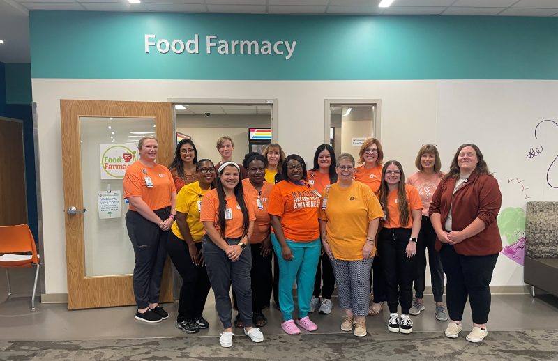 The Food Farmacy standing together and smiling in front of a door labeled "Food Farmacy" at Akron Children's. They are wearing orange shirts and name badges.