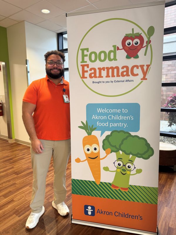 A Food Farmacy team member wears a bright orange shirt and stands next to a banner for the "Food Farmacy" at Akron Children's. The banner features cartoon images of a carrot and broccoli.