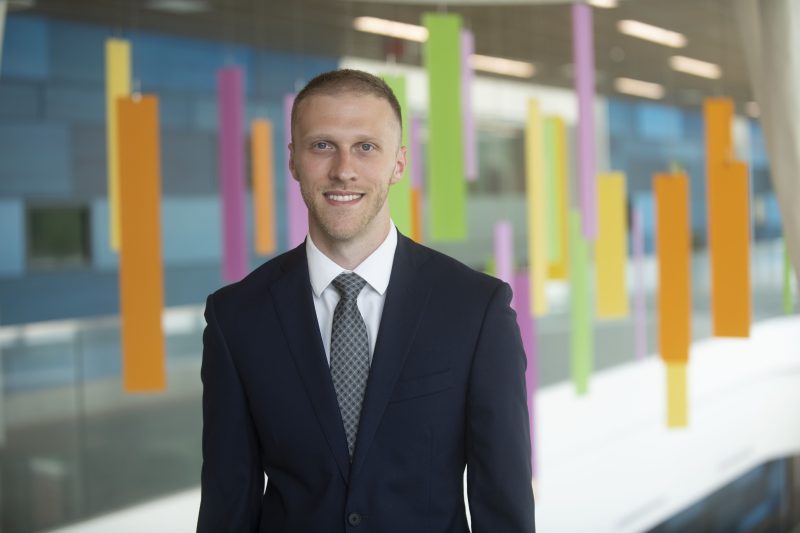 Portrait of Dr. Corey Scherer, gastroenterologist, at Akron Children's. He is standing in a suit and tie smiling at the camera. There are decorative rectangles hanging from the ceiling in the background. They are orange, yellow, green, and pink.