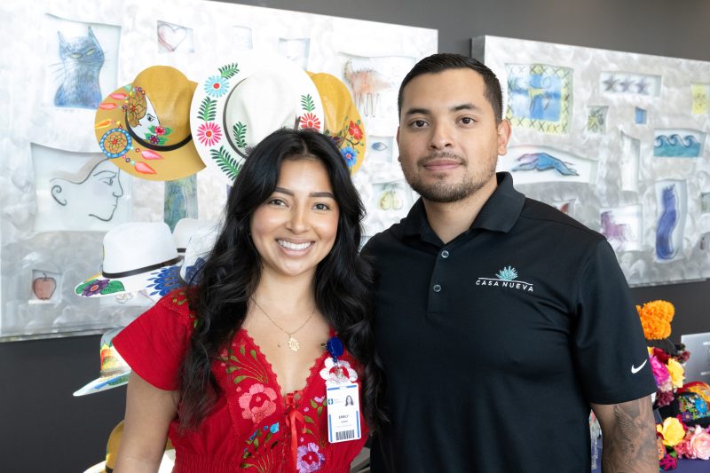 Emely Lemus and another individual standing side by side, smiling, in front of a colorful display of hats and art. One person is wearing a red outfit with embroidered designs and a name badge. The other person is wearing a black polo shirt with the "Casa Nueva" logo. The background includes artistic hat designs and sketches.