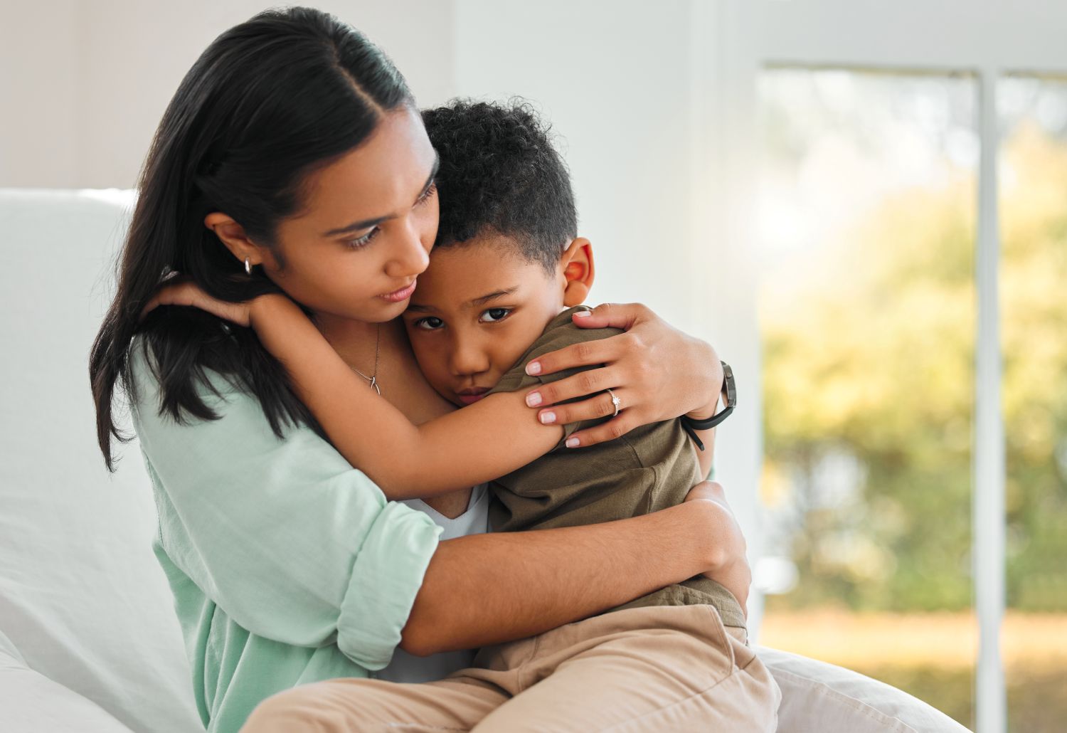 A photograph of a mother comforting her young son. She holds him in her arms while the child looks towards the camera with a worried look. The background seems to be inside with two glass doors behind them.