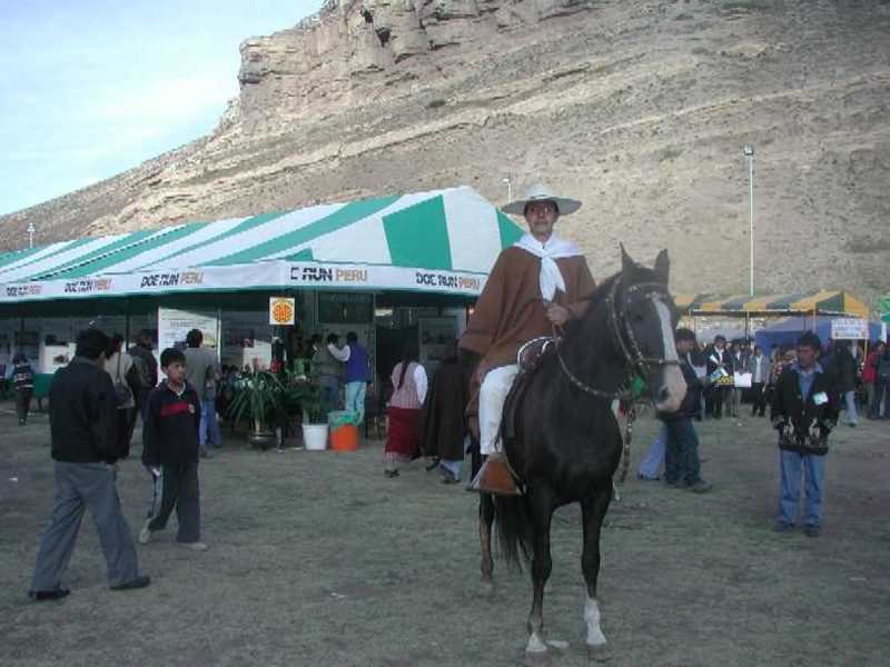 Jesus Gutierrez Castilla is sitting on a horse in front of a pavilion with "DOC RUN PERU" written on it. The scene includes other people walking around and a rocky hillside of Peru in the background. The setting appears to be an outdoor event.