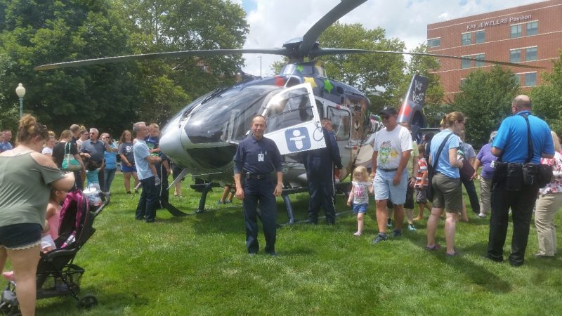 A group of people is gathered around a parked helicopter on a grassy area. Jesus Gutierrez Castilla stands in front of Air Bear, Akron Children's rotor wing aircraft. The helicopter bears a medical insignia. People are engaging with the scene, some taking photos.