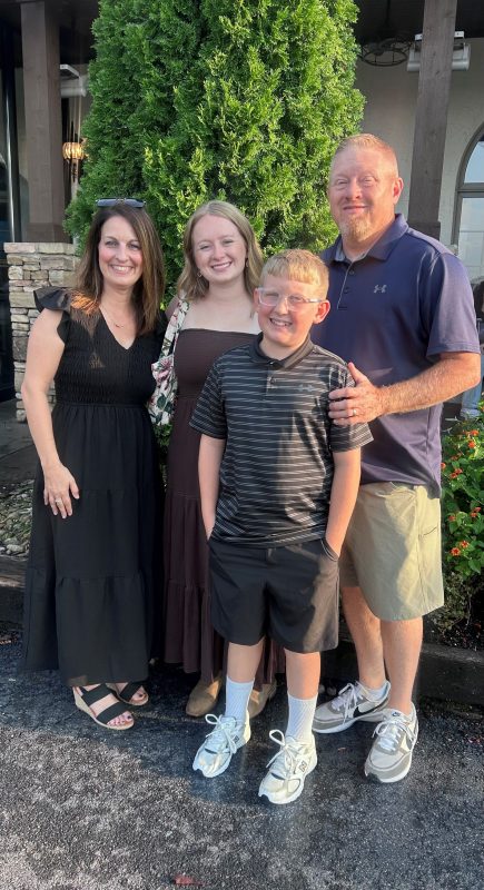 Jacob (Jake) Morrison and his family. On the left is his mother, Antoinette, in a black dress, his sister, Ella, in a brown dress, and his dad, Bob, in a navy polo and khaki shorts. Jake is standing in front of them with his dad's hand on his shoulder. Jake is wearing a black striped polo and black shorts. They are standing in front of some greenery.