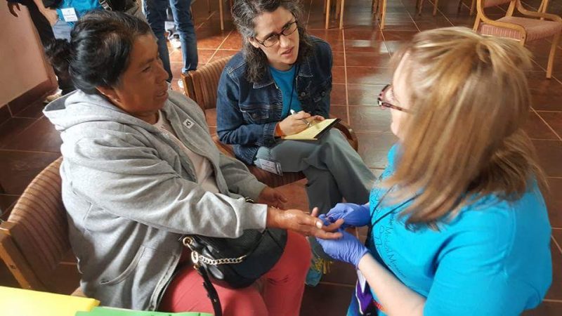Emily Lanier translating between a medical professional and a Hispanic-speaking patient. The nurse is holding the hand of the patient, who is seated and wearing a gray jacket. Emily Lanier, wearing glasses and a denim jacket, is taking notes and translating.