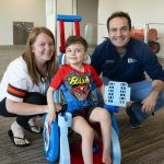 Courtney (left) and Dr. Magoline (right) are crouching down beside Colt (middle) who is sitting in his custom-made toddler mobility trainer (TMT). They are looking towards the camera smiling in a lobby at Akron Children's. Courtney is wearing a Browns jersey, Colt is wearing a Flash t-shirt and shorts, and Dr. Magoline is wearing an Akron Children's navy-blue polo.