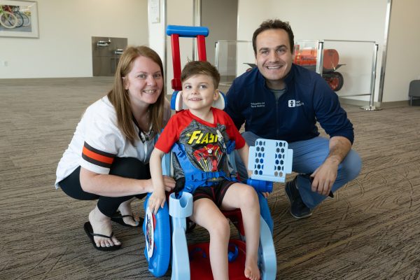 Courtney (left) and Dr. Magoline (right) are crouching down beside Colt (middle) who is sitting in his custom-made toddler mobility trainer (TMT). They are looking towards the camera smiling in a lobby at Akron Children's. Courtney is wearing a Browns jersey, Colt is wearing a Flash t-shirt and shorts, and Dr. Magoline is wearing an Akron Children's navy-blue polo.