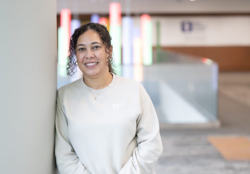 Cassandra (Cassie) Neely leaning against a pillar at Akron Children's, wearing a light-colored Akron Children's branded sweatshirt. The background features colorful vertical light bars and a blurred logo of Akron Children's on the wall.
