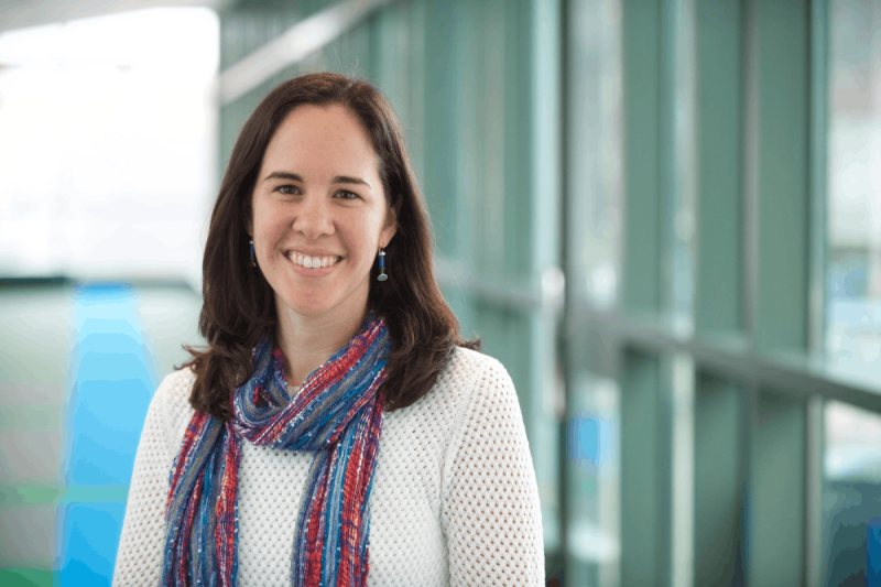 Portrait of Akron Children's pediatrician Hanna Lemerman, MD. She is wearing a white knit sweater with a blue and red scar around her neck. The background appears to be a hallway in the hospital lined with large windows.