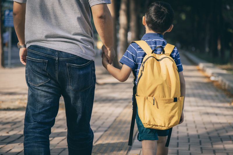The image shows a father and son walking to school on a brick sidewalk. The child is wearing a yellow backpack and blue striped shirt. The father wears a gray shirt and jeans. They are holding hands. The picture is taken from behind them as they walk.