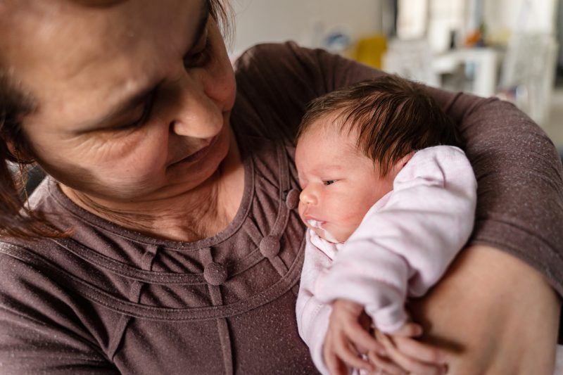 Senior caucasian woman grandmother hold newborn infant girl vomiting milk – baby overfeeding puke milk in hands of her granny at home – new life nursing and growing up concept