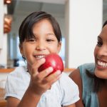 The importance of digestive health to your child’s growth Mom and young daughter smiling as the girl holds a bright, red apple.