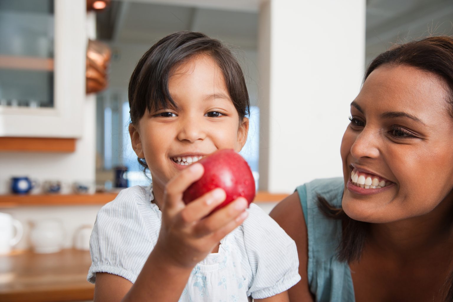 Mom and young daughter smiling as the girl holds a bright, red apple. The background appears to be in a home kitchen.