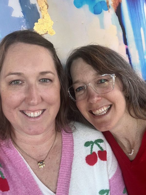 Kristin Selby (left) and Allison Dennison (right) smiling in close-up photo. Kristin is wearing a white sweater with cherries on it, and Allison is wearing a red sweater and clear glasses.