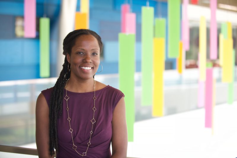 Portrait of gastroenterologist Dr. Christine Carter-Kent. She wears a purple blouse with a long gold necklace. She is smiling at the camera, and the background shows decorative and colorful rectangles hanging from the ceiling.