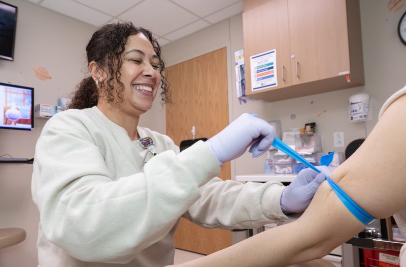 Cassandra (Cassie) smiling and preparing to draw blood from a patient's arm using a tourniquet in an Akron Children's lab. The room has medical posters, a clock, and equipment.